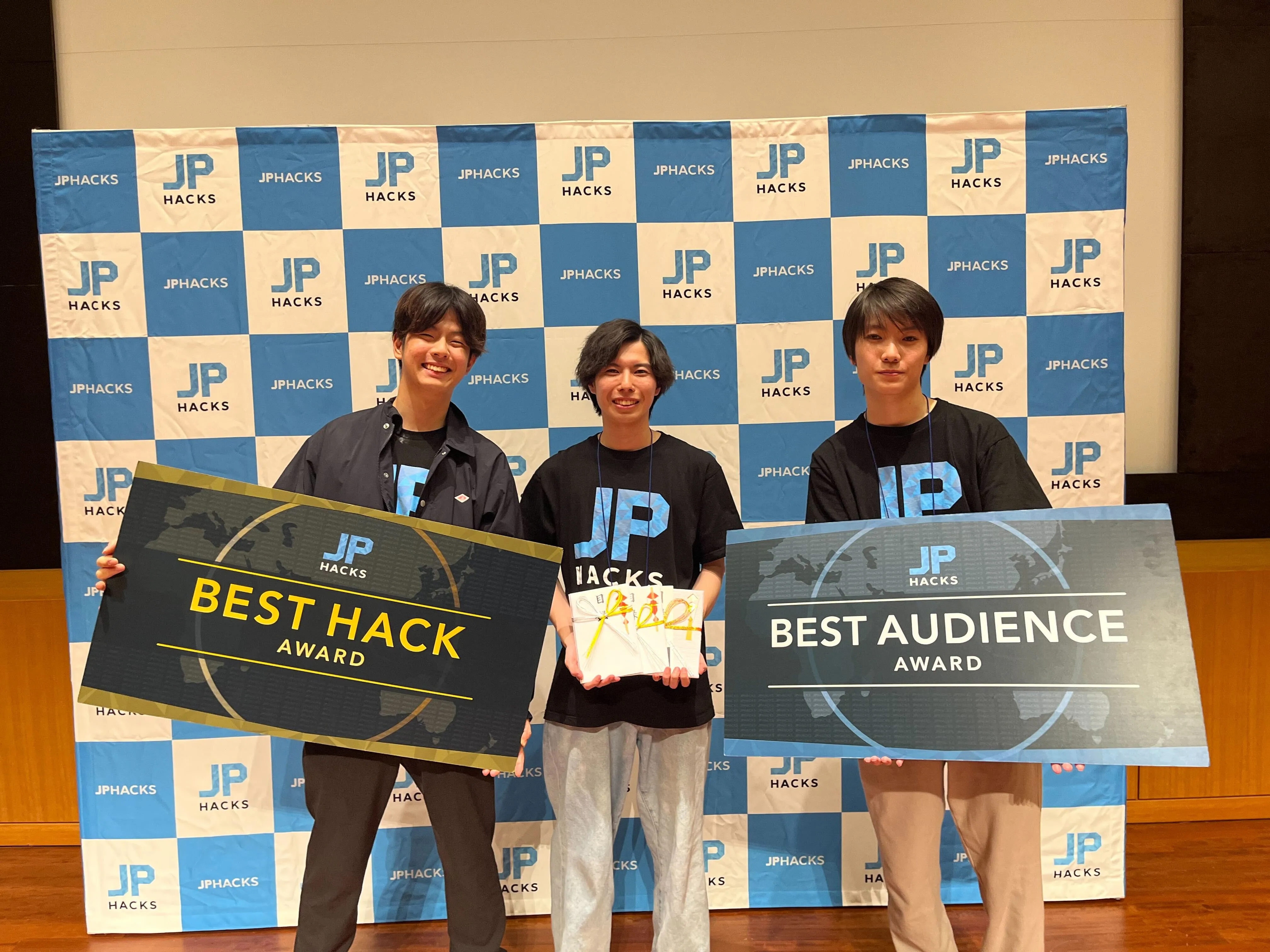集合写真。3人の男性が正面を向いており、左右の男性はJPHacksを優勝したことを表すボードを抱えている。中央の男性は表彰でもらった目録を3つ手に持っている。