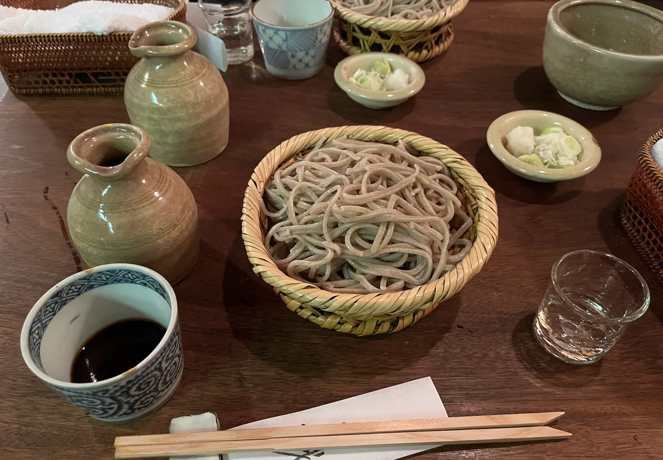蕎麦の写真：中央に丸いざるに乗ったざるそばがあり、左につゆ、右に薬味が乗った皿と、日本酒が注がれたグラスがある。手前には割り箸が置かれている。